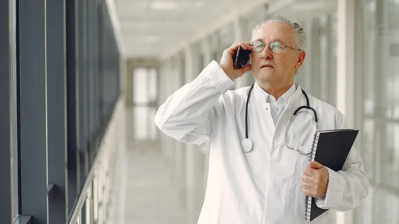 Médico de jaleco branco em pé num corredor claro de clínica, ao telefone, com um caderno debaixo do braço