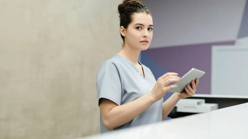 Recepcionista de uniforme cinza-claro atrás do balcão de uma clínica, com um tablet na mão, olhando para a entrada num momento sem ninguém