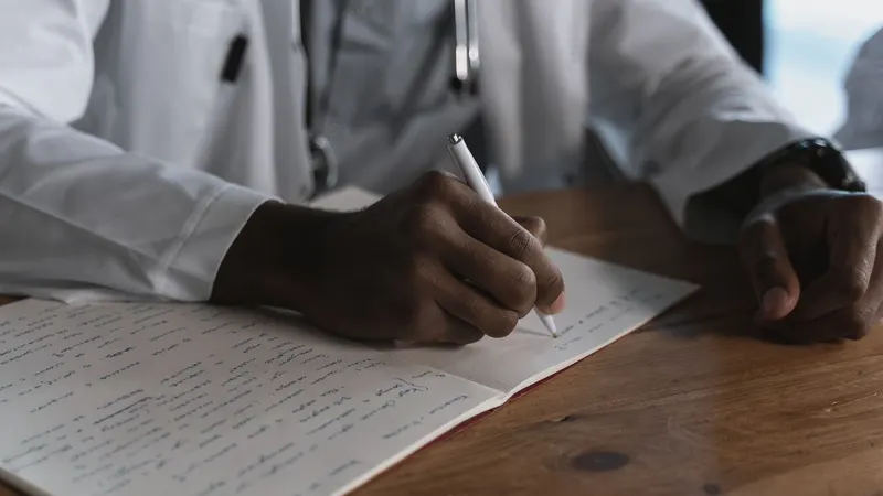 Mãos de um profissional de jaleco branco e estetoscópio escrevendo à caneta num caderno aberto sobre uma mesa de madeira