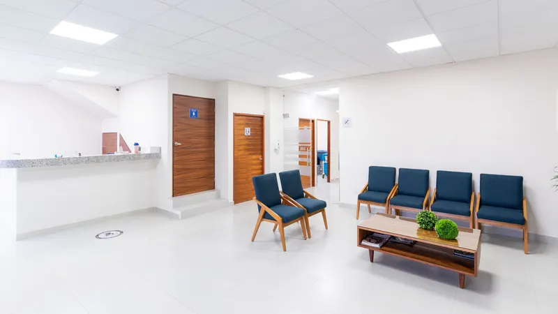 Sala de espera vazia de uma clínica antes de abrir: seis poltronas azuis, uma mesa baixa de madeira com plantas, o balcão de atendimento sem ninguém atrás e, ao fundo, portas fechadas e um corredor que segue para outras salas