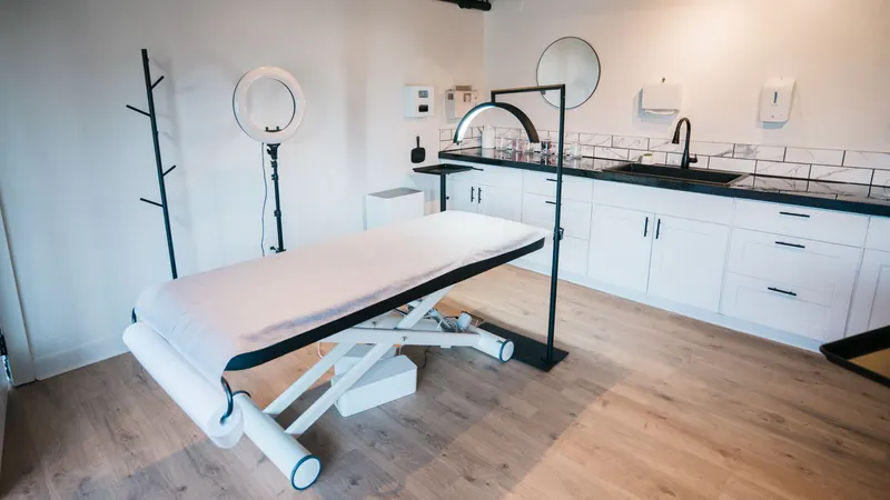 Cabine de clínica de estética montada antes do primeiro horário: maca vazia com lençol claro e rolo de papel na ponta, ring light num tripé, luminária de arco acesa e, ao fundo, a bancada com pia e armários brancos
