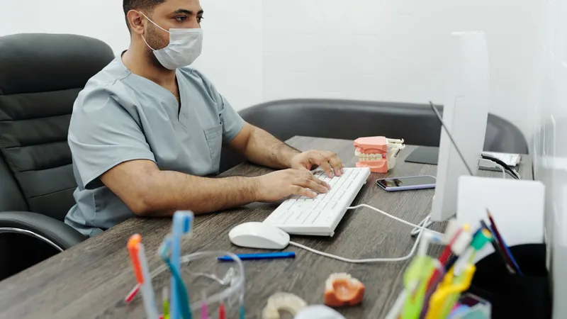 Dentista de máscara e pijama cirúrgico sentado à mesa do consultório, digitando no computador, com um modelo de arcada dentária e um porta-escovas colorido sobre a bancada