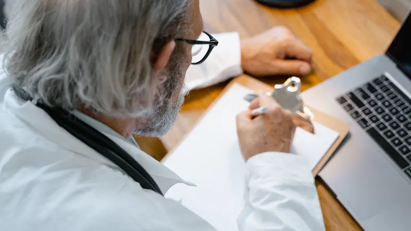 Médico de jaleco branco e estetoscópio, visto por cima do ombro, escrevendo com a caneta numa folha em branco sobre a mesa de madeira, com um laptop aberto de perfil ao lado