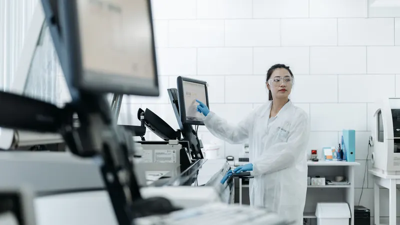 Técnica de jaleco branco, luvas azuis e óculos de proteção opera a tela de um analisador numa bancada de laboratório de azulejo branco, com outros equipamentos de rotina alinhados à frente