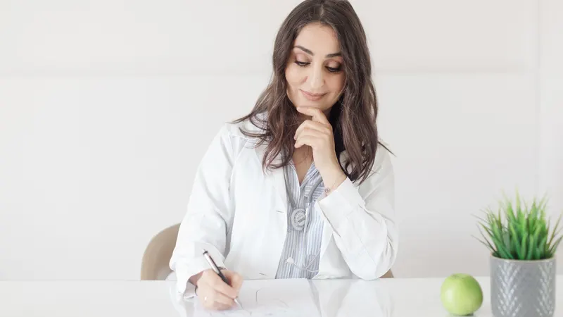 Nutricionista de jaleco branco sentada à mesa clara do consultório, escrevendo numa ficha, com uma maçã verde e um vaso de planta ao lado