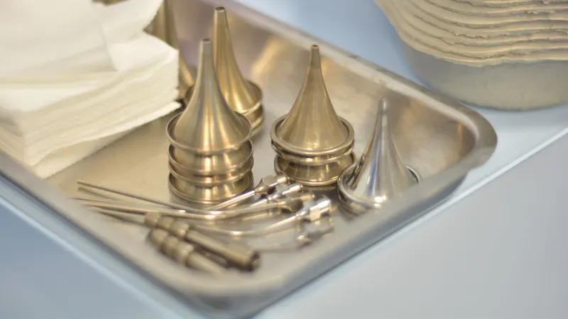 Bandeja metálica montada sobre a bancada clara, com espéculos auriculares de metal em três tamanhos, pinças e curetas finas ao lado de uma pilha de compressas dobradas