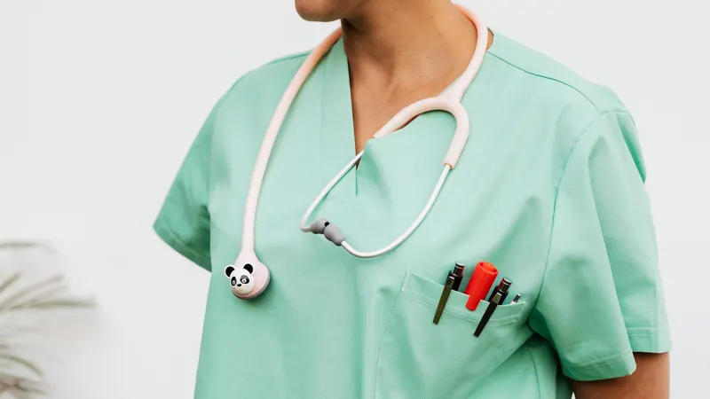 Torso de um profissional de saúde em pijama cirúrgico verde-menta, com um estetoscópio pediátrico de tubo rosa e cápsula em forma de cabeça de panda pendurado no pescoço e três canetas no bolso do peito