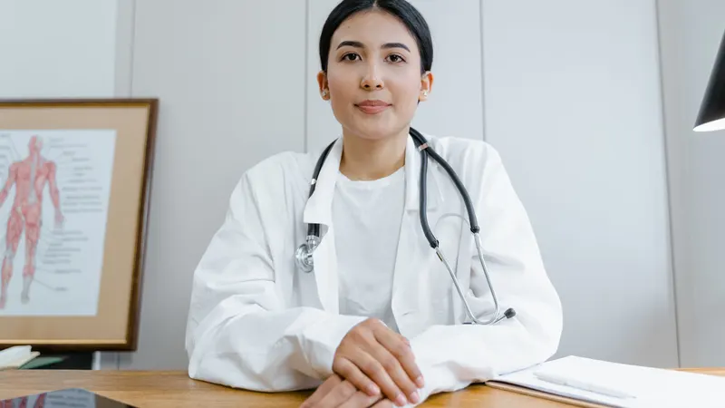 Médica de jaleco branco e estetoscópio sentada à mesa de madeira do consultório, com as mãos entrelaçadas sobre o tampo, um bloco de papel ao lado e uma gravura anatômica emoldurada encostada na parede clara