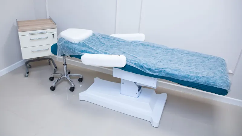 Sala de atendimento vazia antes do horário: maca regulável de base elétrica, com apoio de cabeça e apoios laterais, coberta por um lençol descartável azul, ao lado de um banquinho com rodízios e de um gaveteiro branco