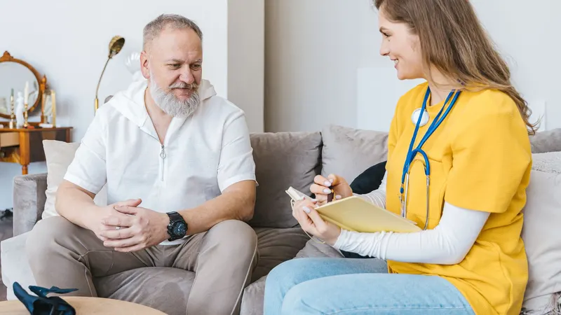 Profissional de saúde sorri e faz anotações enquanto ouve um paciente no sofá do consultório