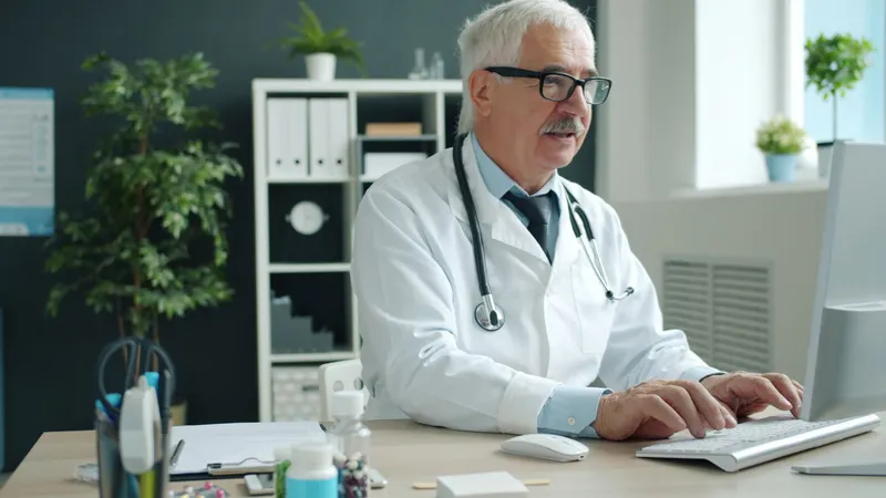 Médico de cabelos brancos, jaleco e estetoscópio digita num teclado à sua mesa, num consultório de parede escura com estante de pastas, plantas e janela ao fundo