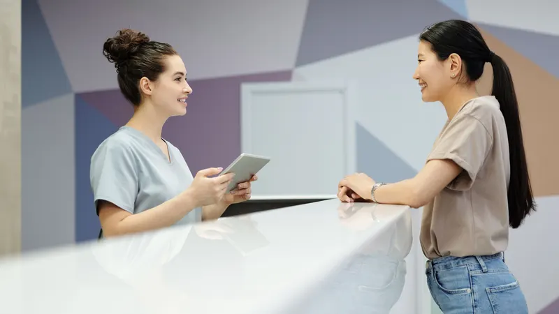 Recepcionista de uniforme azul-claro, com um tablet nas mãos, sorri para uma paciente de camiseta bege apoiada no balcão branco da recepção da clínica