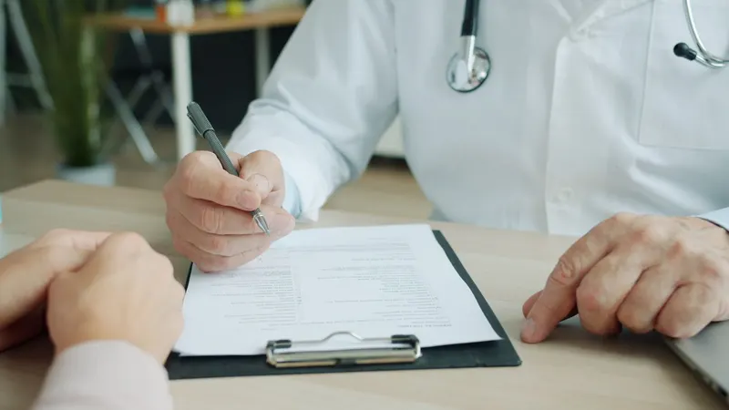 Médico de jaleco branco e estetoscópio escreve com a caneta numa prancheta sobre a mesa clara do consultório, com a mão da pessoa atendida apoiada do outro lado