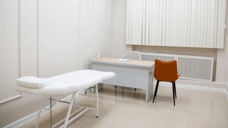 Sala de atendimento vazia e arrumada, com maca forrada de branco, mesa clara, cadeira laranja e persiana filtrando a luz da janela