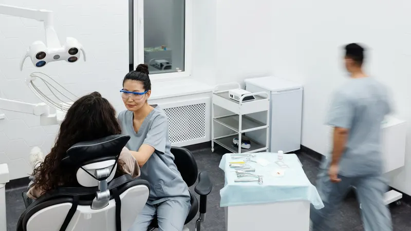 Vista ampla de uma sala odontológica de parede branca em hexágonos, com a dentista de uniforme cinza-azulado sentada ao lado da paciente na cadeira e um colega passando ao fundo