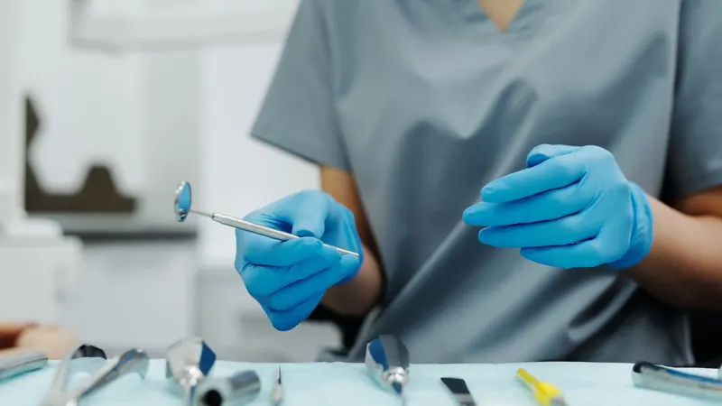Dentista de pijama cirúrgico cinza, enquadrado do pescoço para baixo, segura um espelho bucal com as mãos de luvas azuis sobre a bandeja de instrumentos