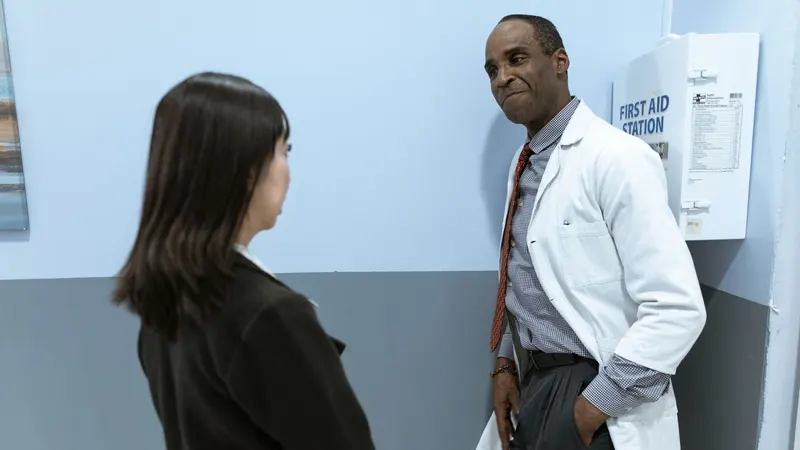 Médico de jaleco branco encostado na parede de um corredor de clínica, conversando de pé com uma pessoa que aparece de costas em primeiro plano