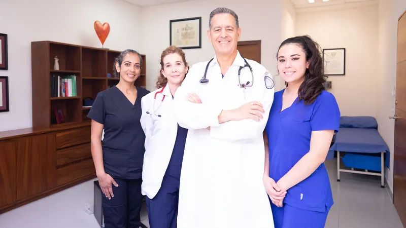 Quatro profissionais de saúde com uniformes diferentes — jaleco branco com estetoscópio, pijama cirúrgico preto e azul — em pé lado a lado, sorrindo, numa sala de clínica de paredes claras com maca ao fundo