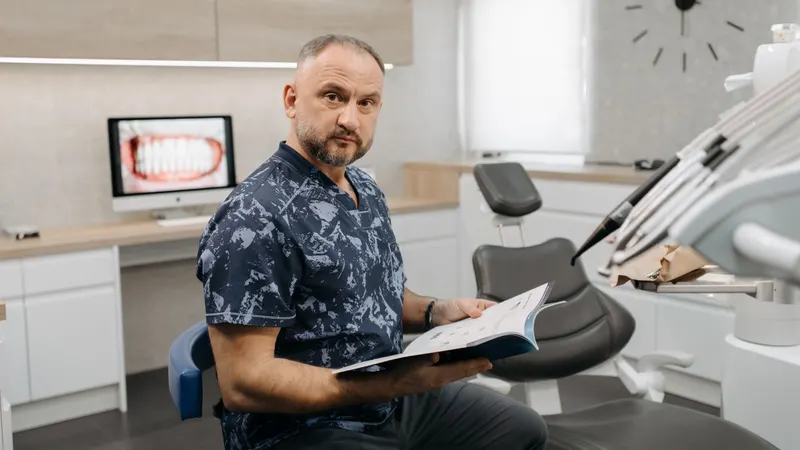 Dentista de camisa estampada, sentado ao lado da cadeira odontológica, segura um caderno aberto e olha para a câmera; ao fundo, a bancada clara e um relógio grande na parede