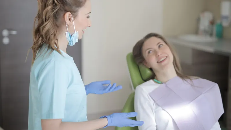 Dentista de uniforme azul-claro, máscara no queixo e luvas, conversando em pé e sorrindo com uma paciente sentada na cadeira odontológica