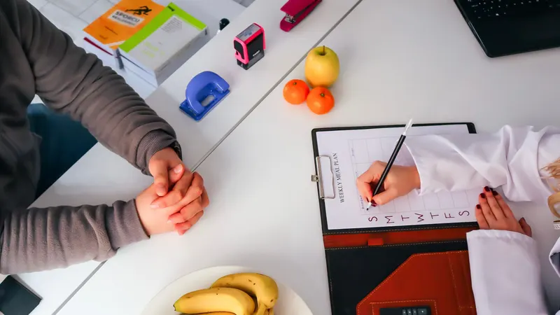Vista de cima de uma consulta de nutrição: a nutricionista de jaleco branco escreve um plano semanal na prancheta enquanto o paciente, de mãos cruzadas, espera do outro lado da mesa com uma fruteira