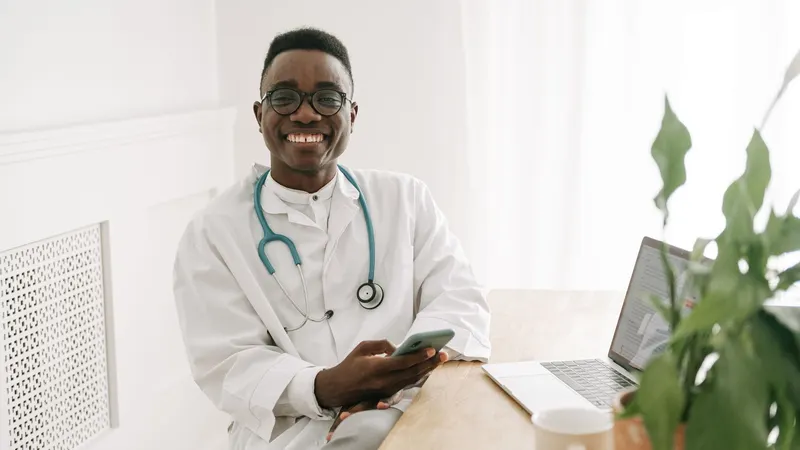 Médico de jaleco branco e estetoscópio, sentado à mesa do consultório com o celular na mão, um laptop aberto ao lado e a luz da janela entrando