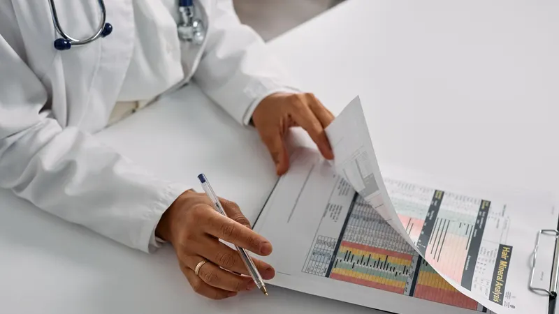 Mãos de profissional de jaleco branco e estetoscópio folheando as páginas de um documento impresso sobre a mesa clara do consultório, caneta na mão