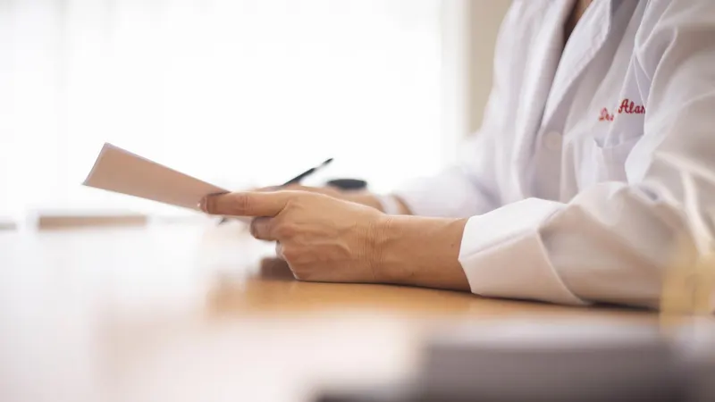 Mãos de uma profissional de jaleco branco segurando um documento sobre a mesa de madeira do consultório, contra a luz da janela