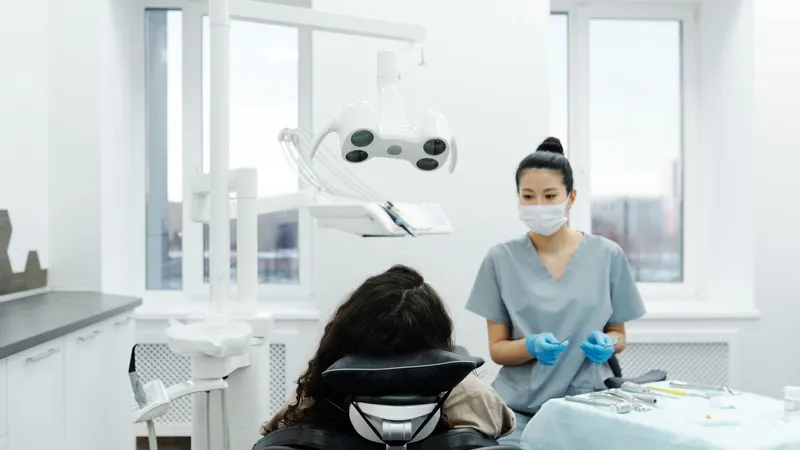 Dentista de máscara e luvas ao lado da cadeira ocupada por uma paciente, numa sala clara de janelas amplas, com o instrumental preparado sobre a bandeja
