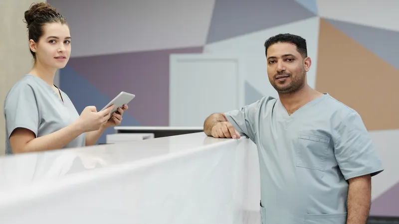 Dois profissionais de uniforme cinza-claro atrás do balcão de recepção de uma clínica, um deles com um tablet na mão, olhando para quem chega