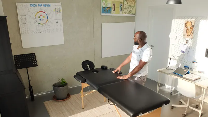 Fisioterapeuta de pé apoia as mãos na maca preta que acaba de abrir, numa sala clara com cartazes anatômicos na parede, planta no chão e mesa branca ao lado